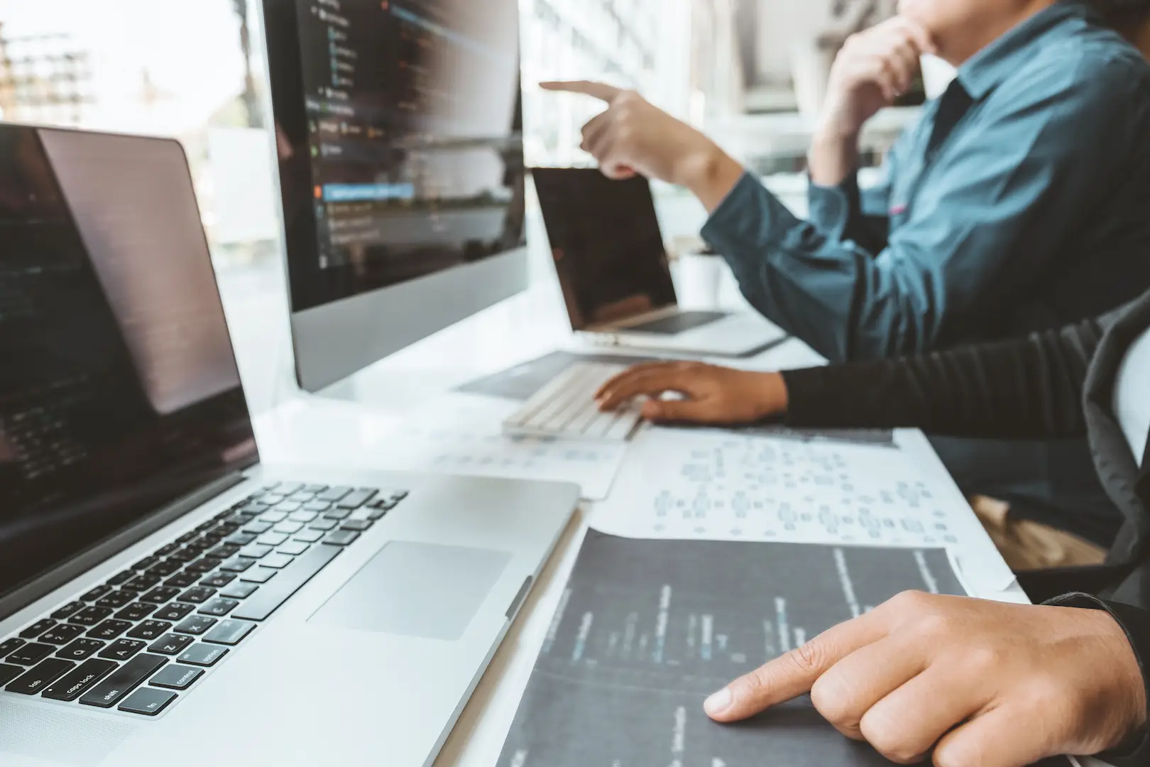 Two people reviewing code on a monitor and printed diagrams at a desk